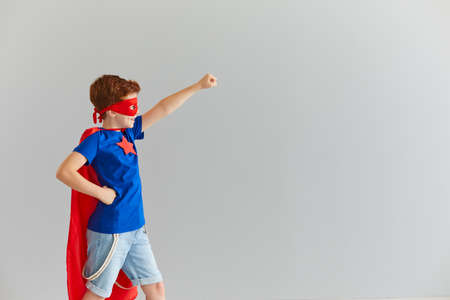 A little smiling boy in a superhero costume with a red cloak and glasses is ready for adventure. Child poses against the background of a gray wall and extends his fist forward. Superhero concept.の写真素材