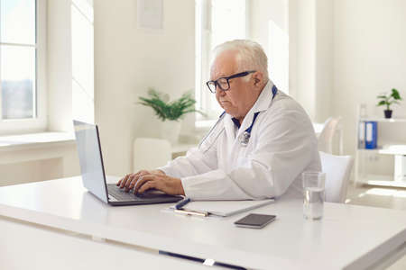 Senior general practitioner working on laptop sitting at desk in hospital office. White-haired doctor giving online consultation to patient on e-health website or entering EHRs into computer systemの写真素材