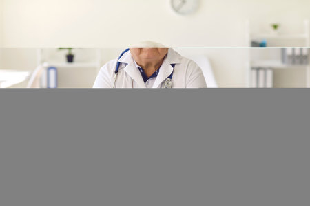 Serious senior male doctor sitting at desk in hospital office and looking at camera, giving online health consultation to patient during video call, or pre-recording medical webinarの写真素材