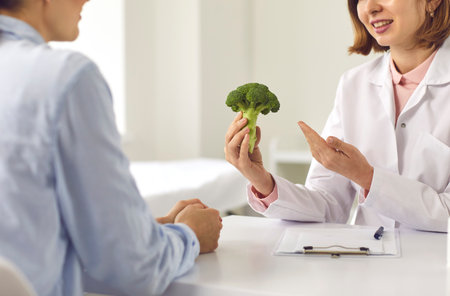 Cropped female doctor, dietitian, nutritionist, food expert or consultant in medical center telling young woman about benefits of healthy diet and recommending eating broccoli and other vegetablesの写真素材