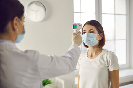 Young woman in medical face mask gets her body temperature checked with non-contact forehead infrared thermometer device before seeing doctor at the hospital. Concept of Covid-19 preventive measuresの写真素材