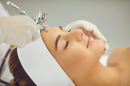 Facial skincare treatment. Young smiling womans face getting oxygen therapy or jet peeling from cosmetologist with special equipment in beauty spa salonの写真素材