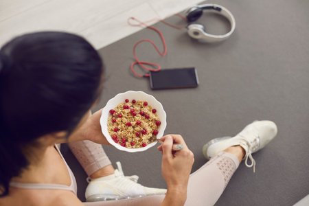 Top view of a bowl of oatmeal with cranberries in the hands of an unknown sports woman. Woman sitting on mat near phone and headphones and eating fresh healthy food after active fitness workout.の写真素材