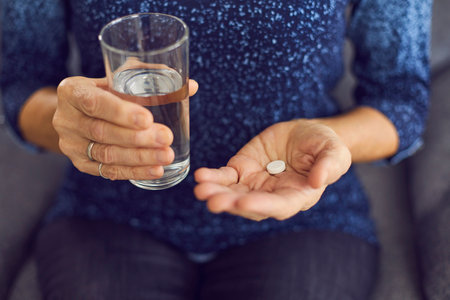 Medication concept. Mature female taking medicine to ease pain, lower blood pressure, or help with stomach ache and indigestion. Closeup of senior womans hands holding glass of water and medical pillの写真素材