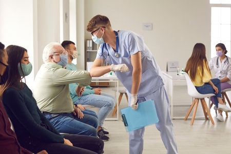 Young man doctor in medical protective face mask bumping elbows with mature senior man patient before vaccination against covid-19 virus during pandemic in medical clinic office. Vaccine against virusの写真素材