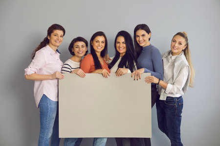 Group team of young smiling women in casual clothing standing and holding big paper sign with mockup blank for text in hands over grey wall background. Women friends having fun together conceptの写真素材