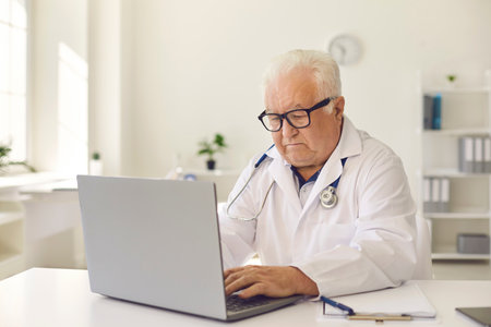 Senior doctor working at desk with laptop in hospital office. Elder practitioner searching for information on the Internet, giving online consultation to patient or entering EHRs into computer systemの写真素材