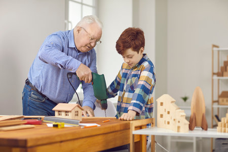 Grandfather and teen grandson working with wood and building toy houses together. Senior man teaches...