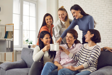Group of happy young women having fun and enjoying time together. Smiling friends or colleagues sitting on sofa at home or in office, laughing at funny joke, clinking cups and drinking coffee or teaの写真素材