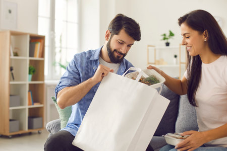 Man and a woman unpack a package of food delivered to them by the delivery service. Couple pulls out food from white package with place for text or logo. Takeaway delivery and food concept.の写真素材