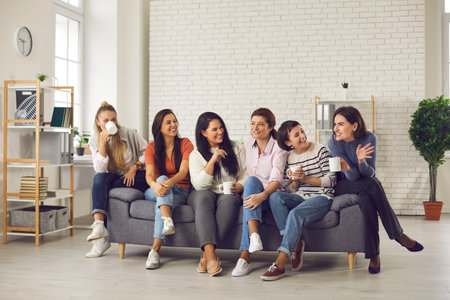 Cheerful friends sitting on sofa at home, enjoying cup of good coffee and nice casual chat together....