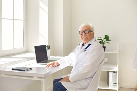 Positive senior professional doctor in uniform sitting with laptop in medical office and looking at camera before online videocall online consultation with patient. Telemedicine and medicare conceptの写真素材
