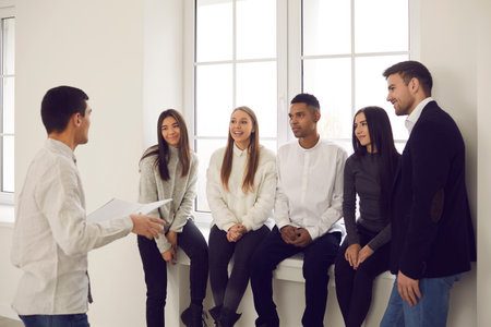 Team of happy young diverse people sitting on windowsill in corporate group meeting, listening to...