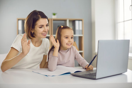 Family staying at home and E-learning via interactive platform. Happy smiling mother and child waving hands at laptop computer, greeting school teacher or private tutor in online lesson via video callの写真素材