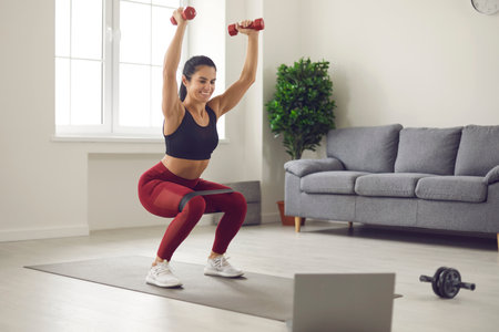 Home online workout in front of a laptop with dumbbells and sports rubber bands. Woman trains squatting and lifting dumbbells up watching online broadcast of fitness exercises Home fitness concept.の写真素材