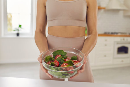 Cropped image of unknown fitness woman holding glass bowl with fresh green basil, cherry tomato and sesame seeds standing at home in the kitchen. Concept of healthy eating, fitness and weight loss.の写真素材