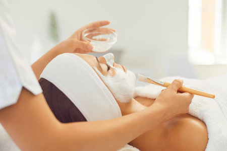 Professional cosmetologist applies a wellness moisturizing cream to a womans face at the spa. Serene young woman is lying on table and enjoying treatment. Beautician uses a cosmetic brush.の写真素材
