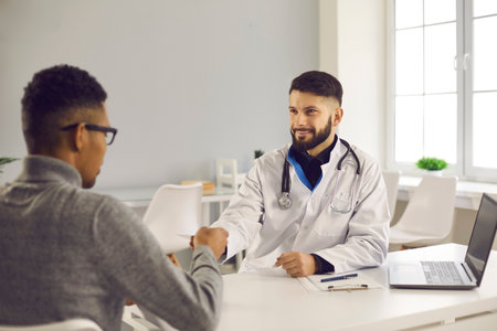 Smiling man doctor and african american man patient shaking hands after successful checkup consultation in medical clinic office. Healthcare, medicare, communication with doctor conceptの写真素材