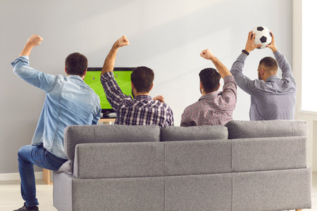 Four excited men friends sitting backwards on sofa at home and watching emotionally soccer football match on television together with hands raised. Leisure, hobby, entertainment for male companyの写真素材