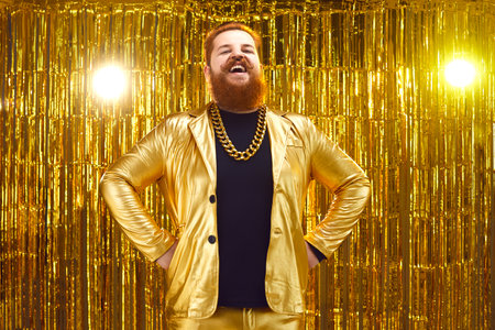 Portrait of funny happy smiling guy on stage with golden background. Content chubby bearded young...