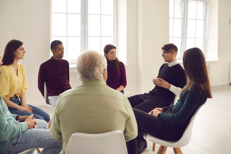 Diverse people listening to therapist sitting in circle in group therapy session. Patients talking, sharing concerns, coping with anxiety, dealing with psychological problems and insecurities togetherの写真素材