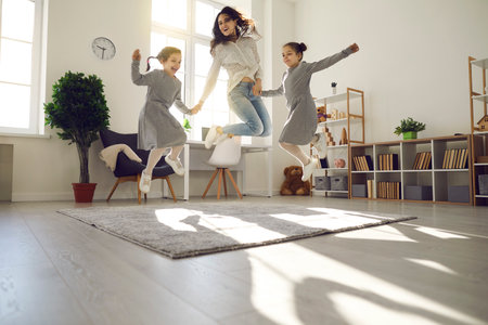Happy family having fun at home. Overjoyed young woman and children enjoying free time and playing together. Excited mother and daughters jumping and laughing in cozy sunlit living-room of modern flatの写真素材