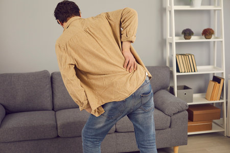 Back view of man walking towards sofa, feeling intense pain and touching his lower back. Backache,...