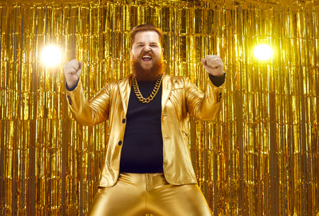 Happy crazy funky uninhibited ginger young man wearing funny golden suit and extravagant gold chain dancing at disco party, celebrating success, having fun, cheering, fist pumping and shouting for joyの写真素材
