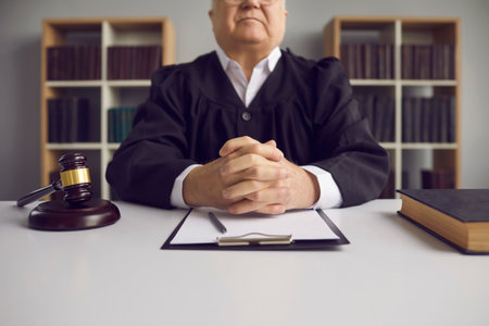 Serious fair incorruptible old male judge at table with gavel and case papers in courtroom. Honest wise senior lawyer in uniform sitting with clasped hands at desk. Concept of court of law and justiceの写真素材
