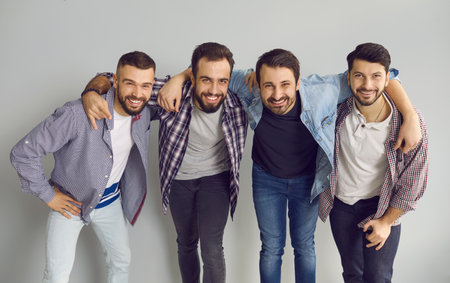 Portrait of happy friends standing with arms around each others shoulders in studio with light gray background. Group of young bearded men hugging, smiling, looking at camera and having fun togetherの写真素材