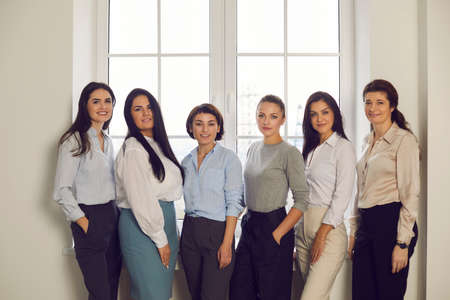 Group portrait of successful confident stylish women in their 20s and 30s standing together by office window. Professional team of happy young company workers or business partners smiling at cameraの写真素材