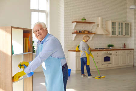 Happy senior couple cleaning their house. Mature husband helping his wife to tidy up at home. Smiling elderly man dusting the shelves while the woman is mopping the floor in their new apartmentの写真素材
