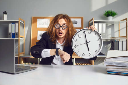 Employee working under pressure. Funny stressed young office worker with messy long hair holding clock, looking at wristwatch and panicking over missed deadline or being terribly late for work meetingの写真素材