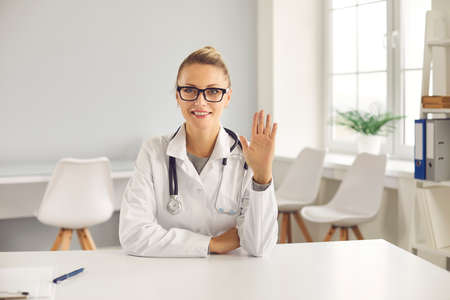 Telemedicine, telehealth, online medicare concept. Young positive blonde female doctor nurse therapist greeting virtual patient with hand during online meeting and consultation from medical clinicの写真素材