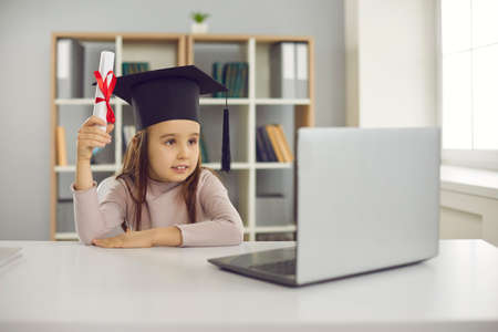 Small cute girl pupil in graduate bonet sitting near laptop and holding diploma in hand during...