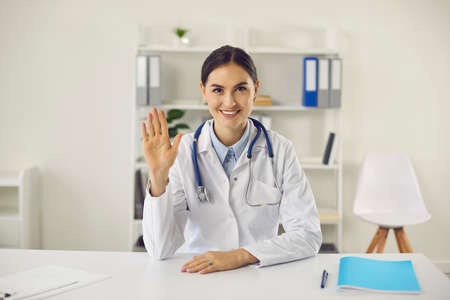 Telemedicine and online medicare concept. Young smiling woman doctor therapist sitting in clinic and greeting patient online with hand during online consultationの写真素材