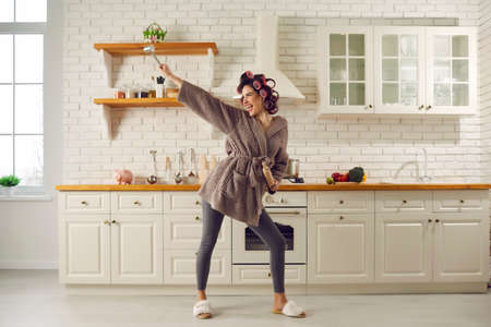 Happy housewife having fun while making breakfast. Beautiful young woman in hair curlers, bathrobe, leggings and house slippers holding cooking utensils, dancing and singing songs in spacious kitchenの写真素材