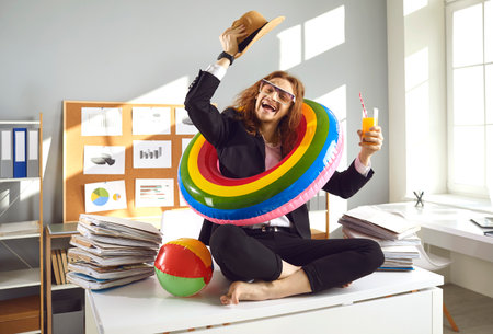 Happy office worker ready for summer holiday. Smiling corporate employee with beach sun hat, sunglasses and colorful inflatable swim ring sitting on desk, sipping drink and dreaming of future vacationの写真素材