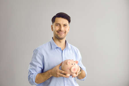 Portrait of happy handsome young man in blue shirt holding pink piggy bank standing against light gray studio background. Concept of finance, banking, business, earning money and saving upの写真素材