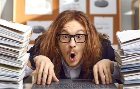 Funny shocked nerdy young accountant sitting at office desk with laptop and folders with paperwork and looking open mouthed at camera with surprised expression on face impressed by crazy money amountの写真素材