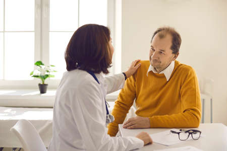 Kind smiling doctor and patient looking at each other. Caring understanding medical worker touches mans shoulder supporting him. Healthcare for senior people, trust, professional consultation conceptの写真素材