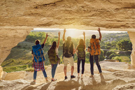 Great achievement. Back view of group of tourists with backpacks standing arms raised at cave entrance on mountain top admiring magnificent nature. Young hikers traveling during summer vacationの写真素材