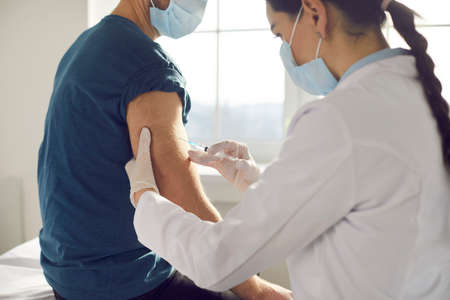 Female medical worker in uniform and face mask injecting vaccine and making vaccination for man...