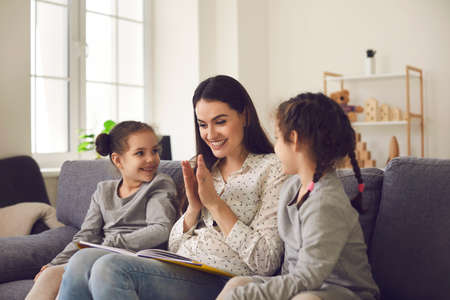 Caring smiling mom reading a book with her two little daughters sitting on the couch at home. Mom or...