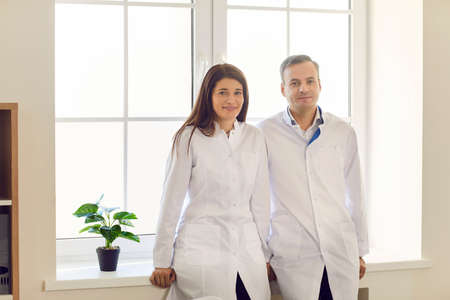 Portrait of two happy male and female doctors in medical uniform standing by the window and smiling looking at camera. Concept of professional medicine and a strong medical team.の写真素材