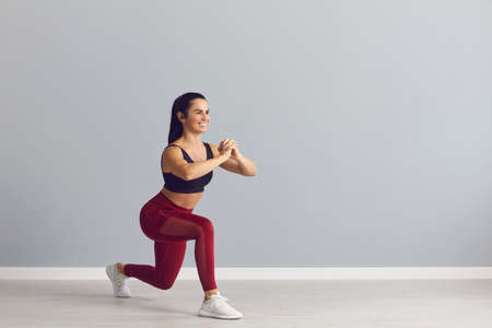 Smiling fitness trainer showing how to do fitness exercise. Happy fit young woman in sports bra and leggings doing forward lunges holding hands together in front of chest during workout at the gymの写真素材