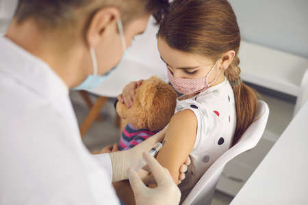Little girl in protective medical face mask sitting with toy and getting vaccination injection with...