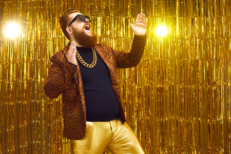 Excited rich red-haired young man in neck chain and cool glasses having fun at glamour night club disco party. Happy fashionable bearded showman singing and dancing on stage with golden backgroundの写真素材
