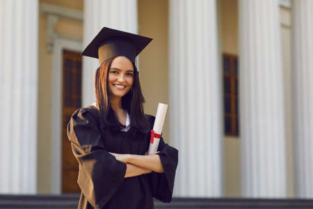 Portrait of confident female student with diploma in hands on university background. Girl is dressed...