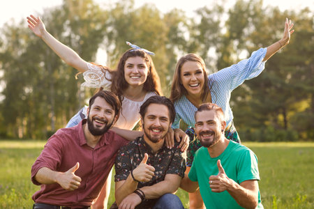 Company of content friends smiling and looking at camera while standing on green meadow in park